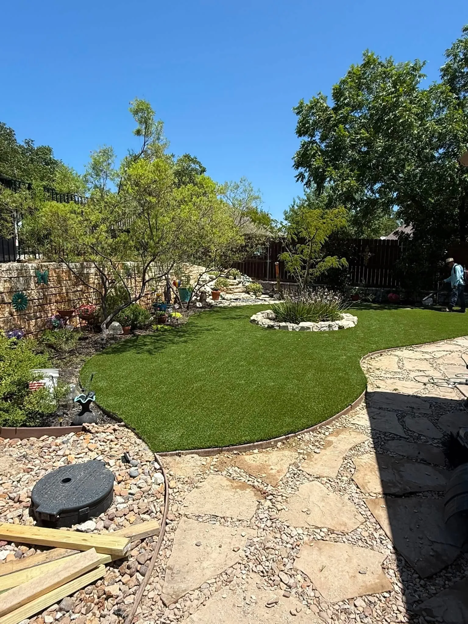 Finished curved artificial turf lawn in a Pecan Plantation backyard, bordered by a stone retaining wall, a rock planting bed, and a flagstone patio.