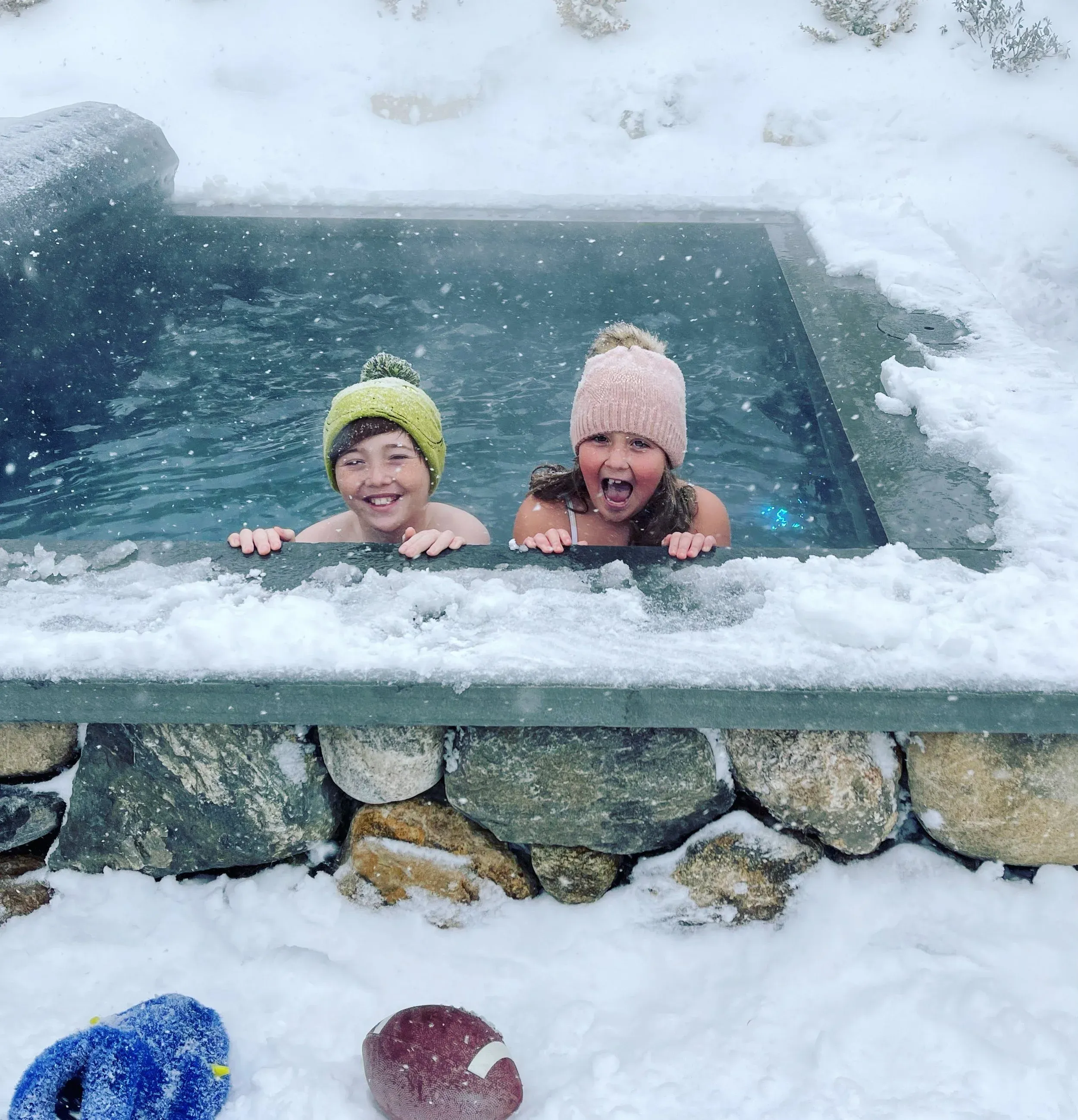 Two kids grinning in a heated Soake plunge pool during a snowstorm